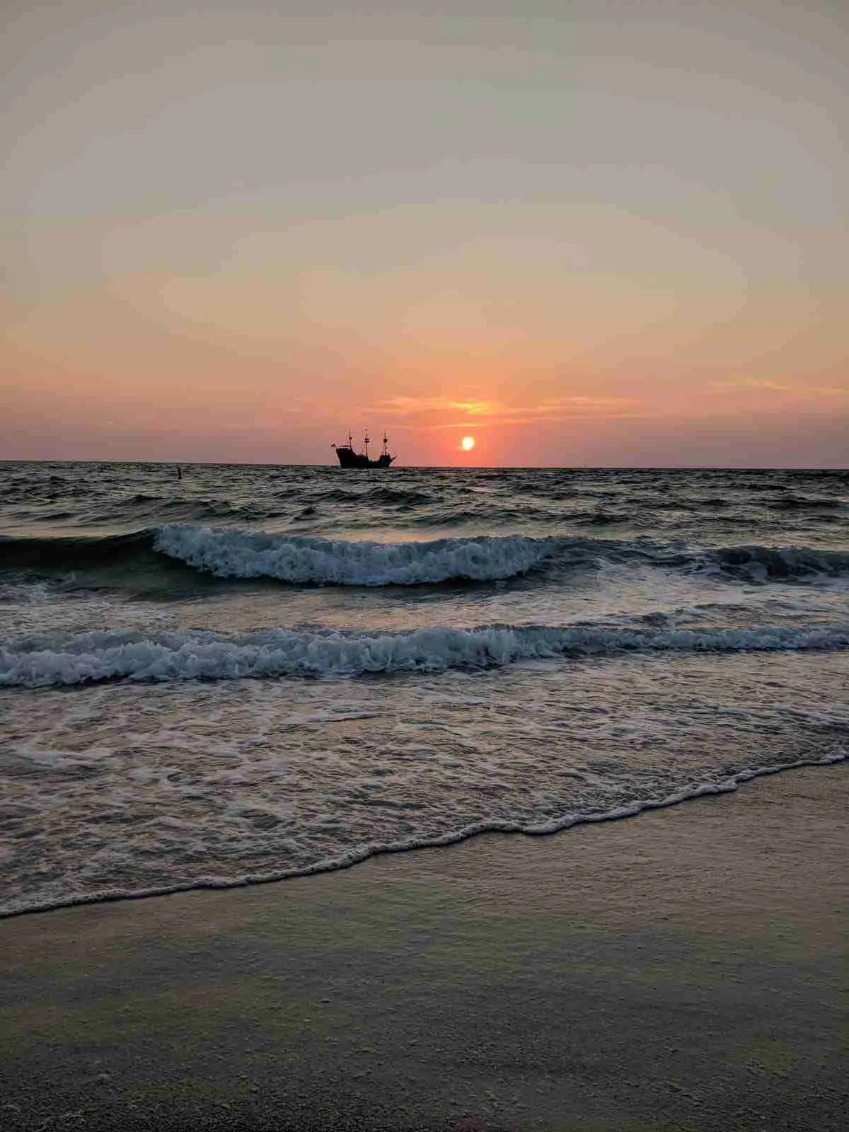 Puesta de sol en la playa con olas y la silueta de un barco de vela antiguo en el horizonte