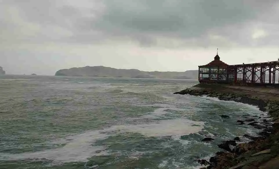 Mar agitado junto a un muelle con un edificio de madera y la costa al fondo