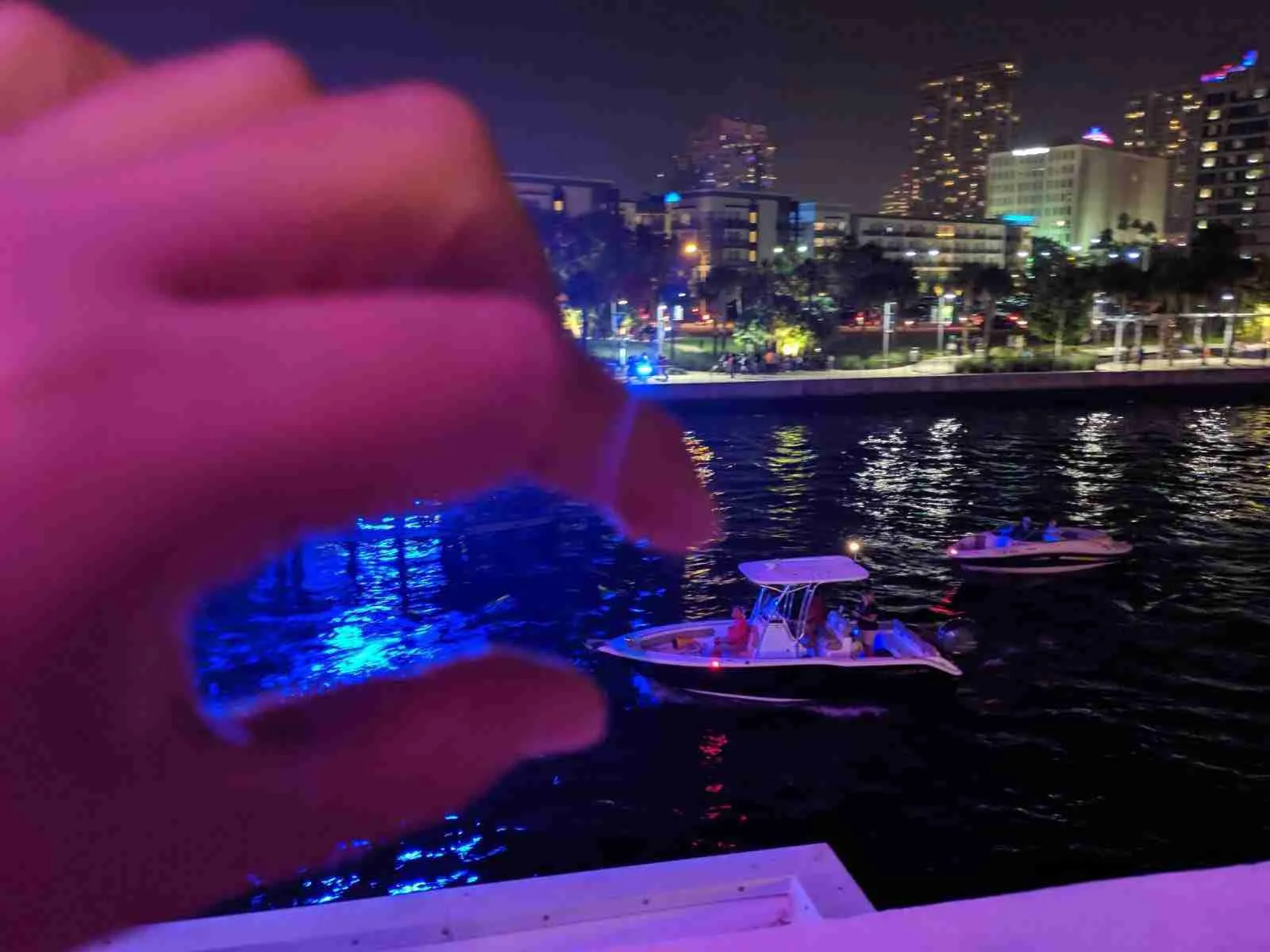 Mano sosteniendo una luz azul frente al paseo marítimo iluminado de noche y una lancha