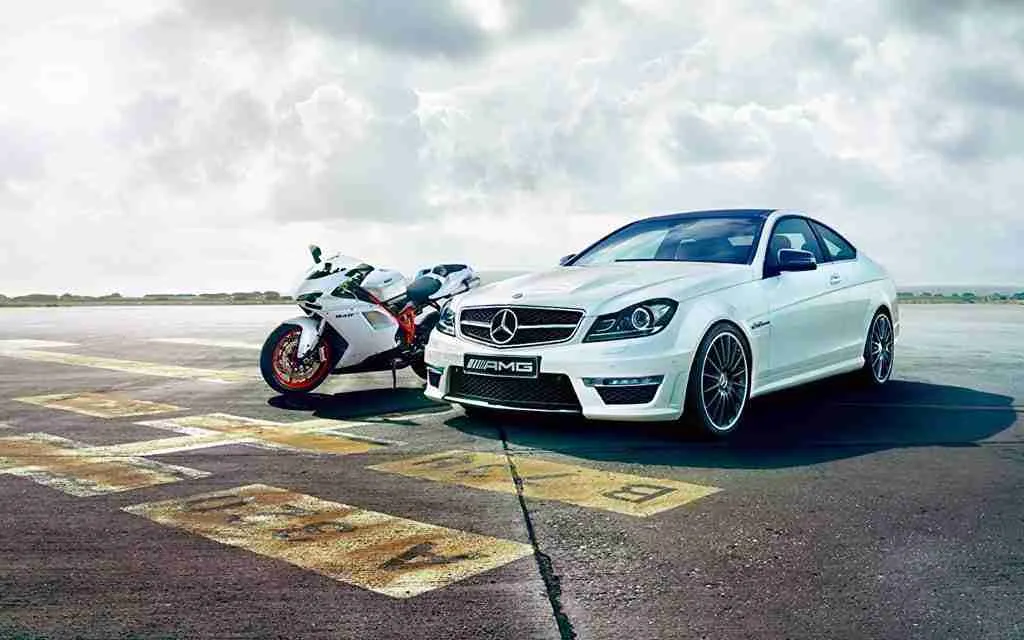 Un coche deportivo blanco y una moto blanca aparcados en una pista bajo un cielo nublado