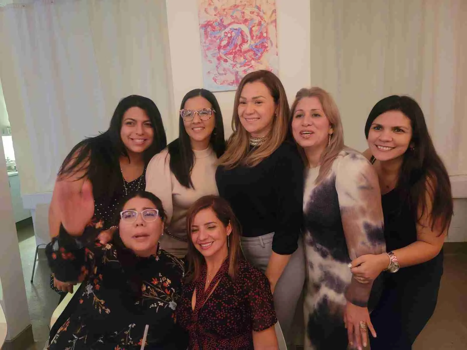 Siete mujeres sonriendo juntas en un salón con un cuadro de colores en la pared
