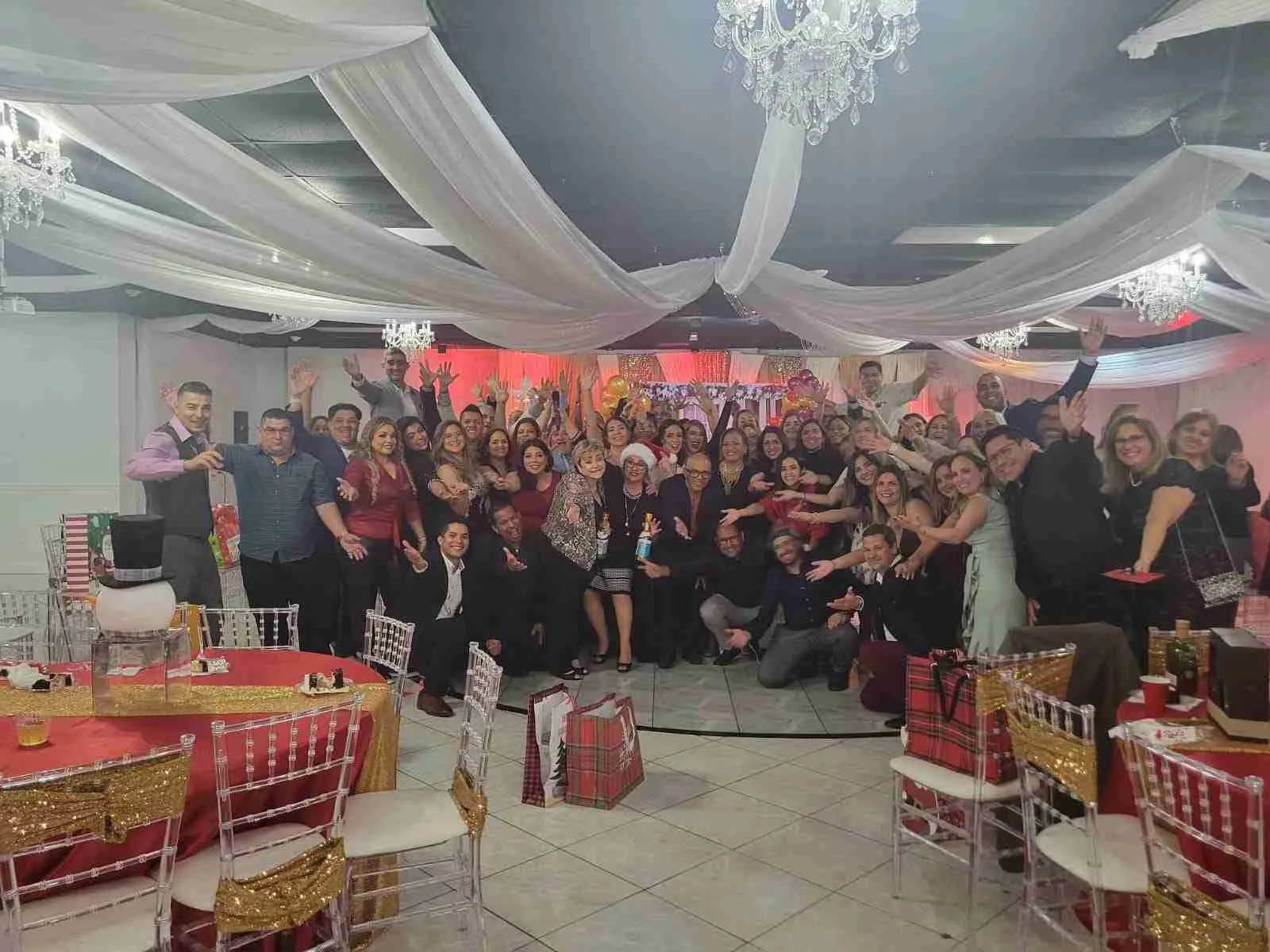 Gran grupo celebrando con los brazos en alto en un salón decorado con telas blancas y lámparas de cristal