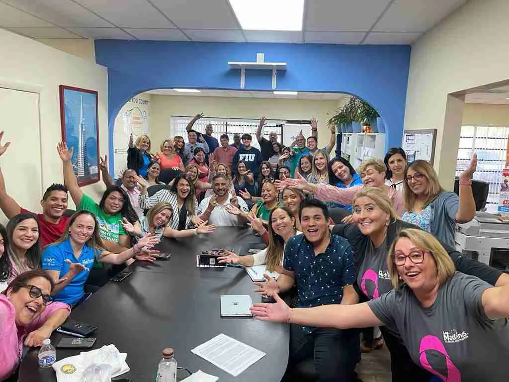 Equipo numeroso saludando a la cámara alrededor de una mesa de reuniones