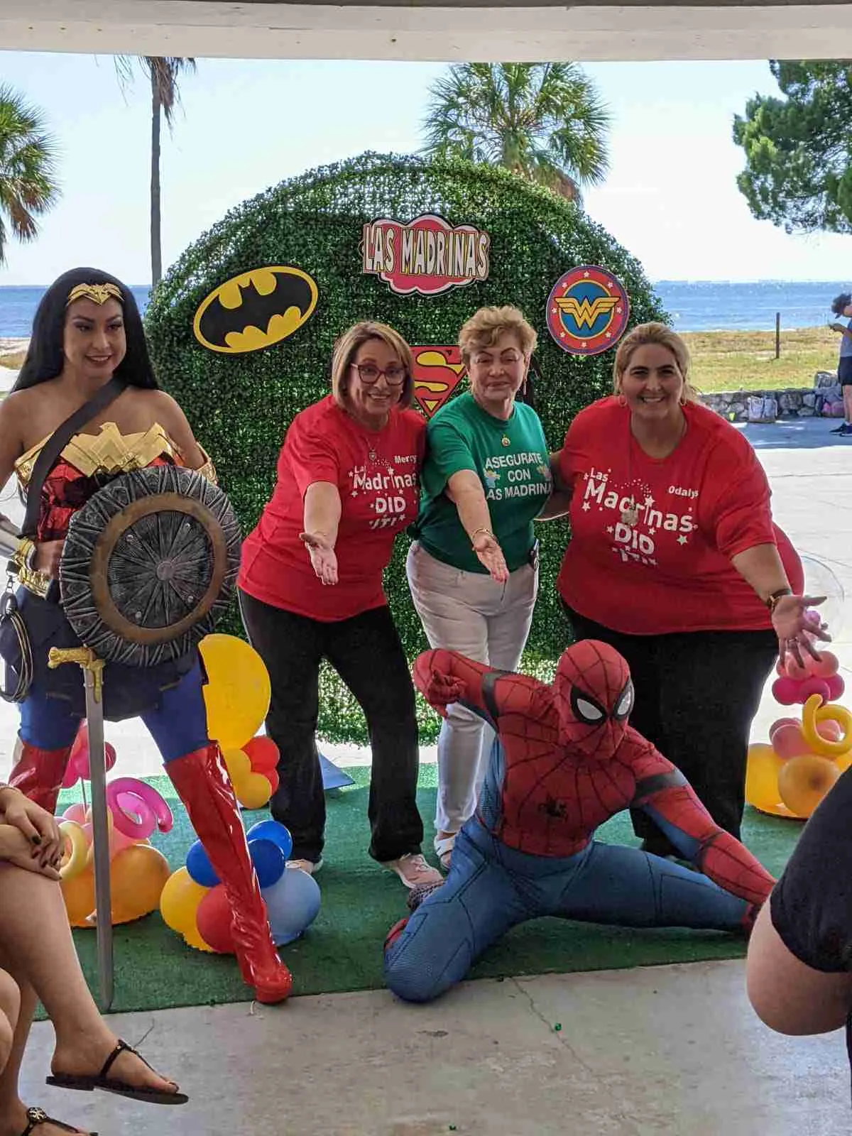 Tres mujeres con camisetas de Las Madrinas posando con personas disfrazadas de Mujer Maravilla y Spider-Man